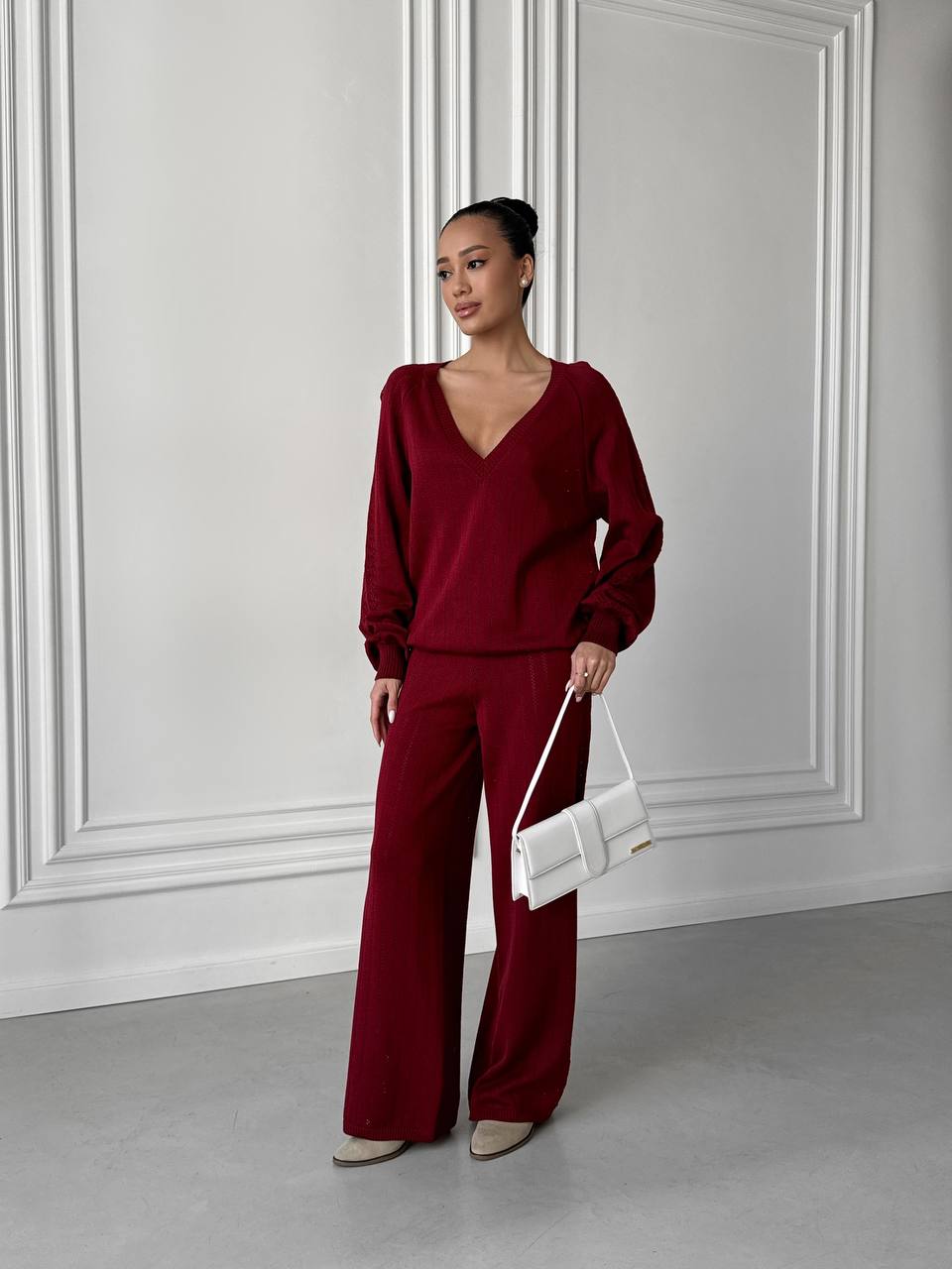 Woman in burgundy outfit holding a white handbag against a gray wall.