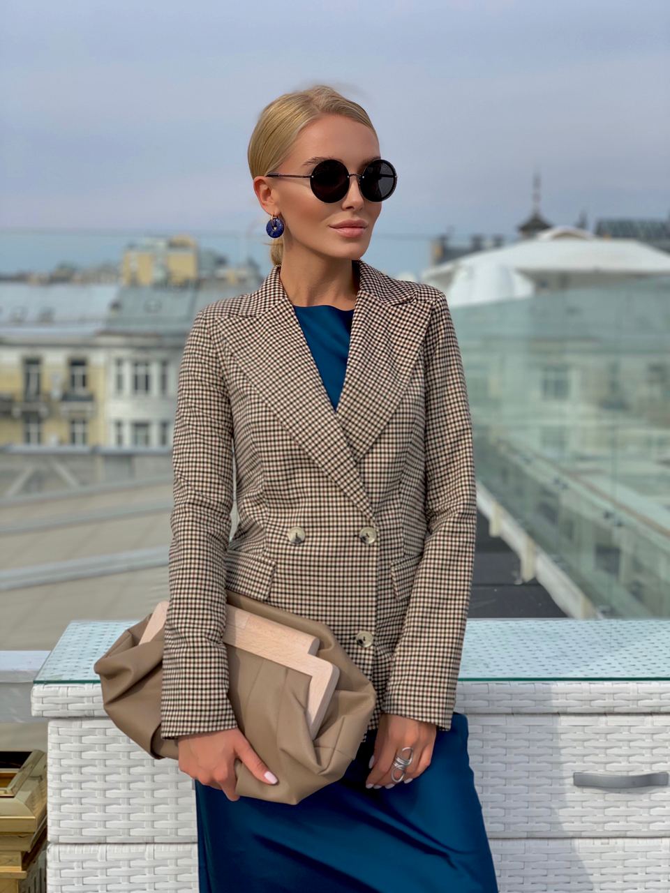 Woman in a plaid blazer and sunglasses standing on a rooftop with cityscape background