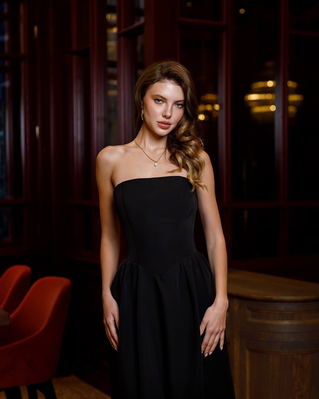 Woman in a black strapless dress standing in a dimly lit room with red chairs.