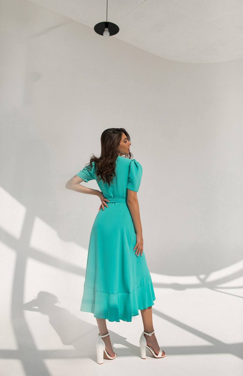 Woman in a turquoise dress standing in a well-lit room with white walls and shadows on the floor.