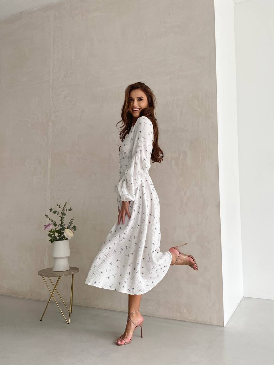 Woman in a white dress standing in a minimalistic room with a vase of flowers on a small table.