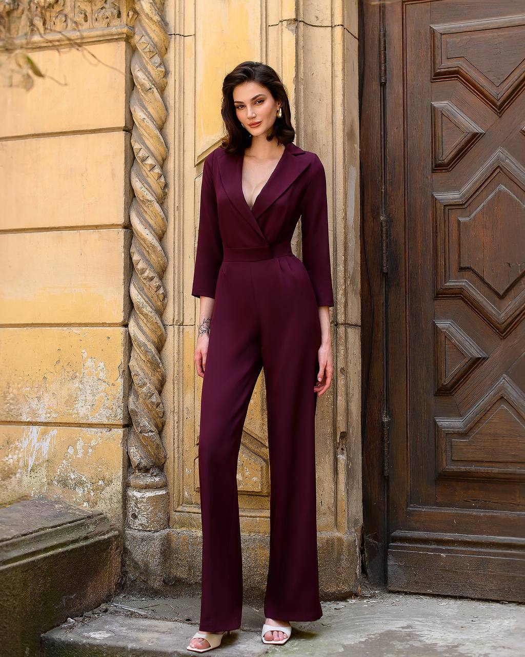 Woman in a deep purple jumpsuit standing in front of an ornate door.
