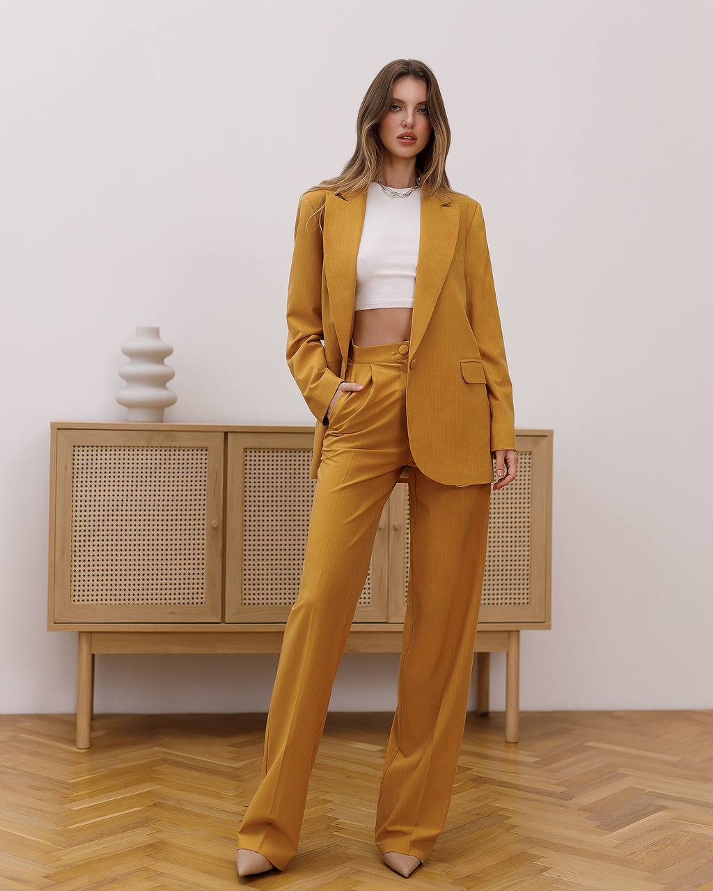Woman wearing a mustard yellow suit standing in a room with wooden flooring and a light-colored wall.