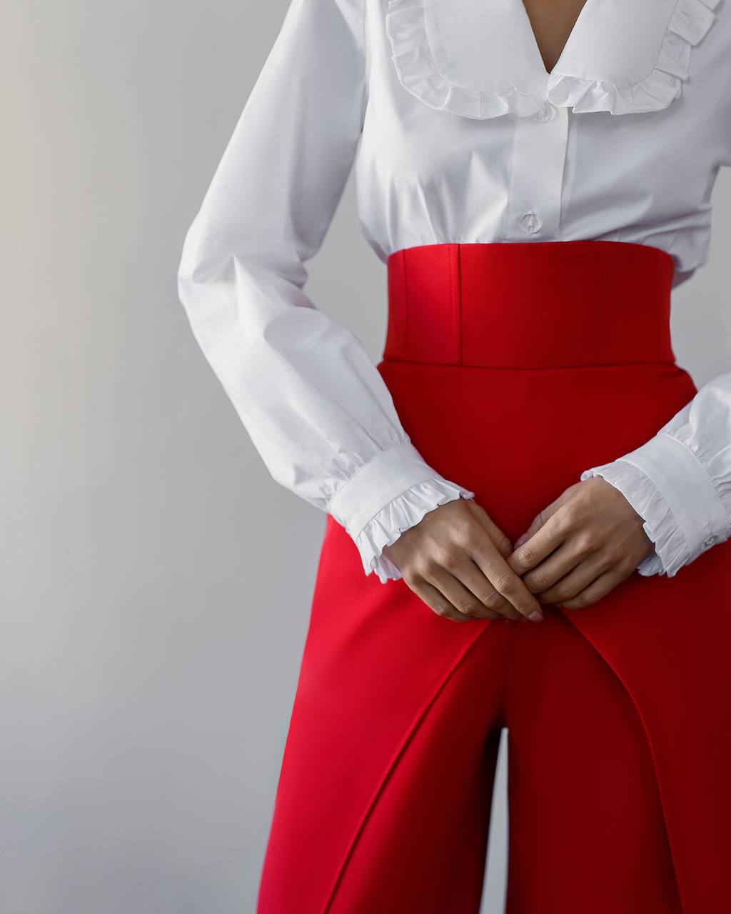 White blouse with ruffled details and red high-waisted pants on a gray background