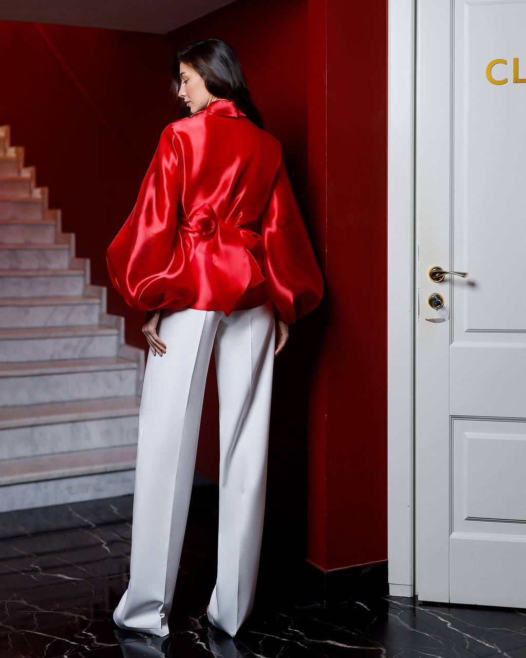 Person wearing a red satin jacket and white pants in a room with a red wall and white door.