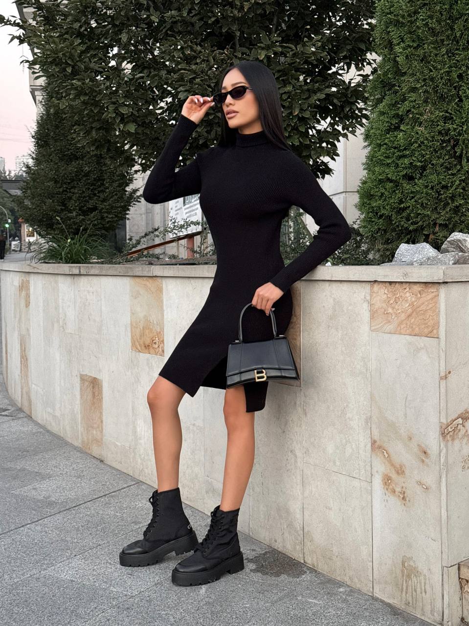 Woman in a black dress and boots holding a black handbag against a stone wall.