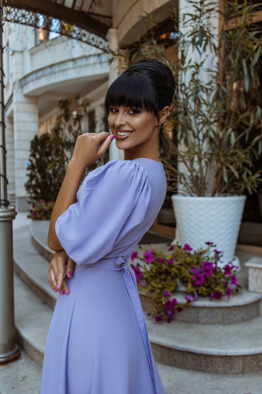 Woman in a lavender dress posing outdoors with flowers and a building in the background