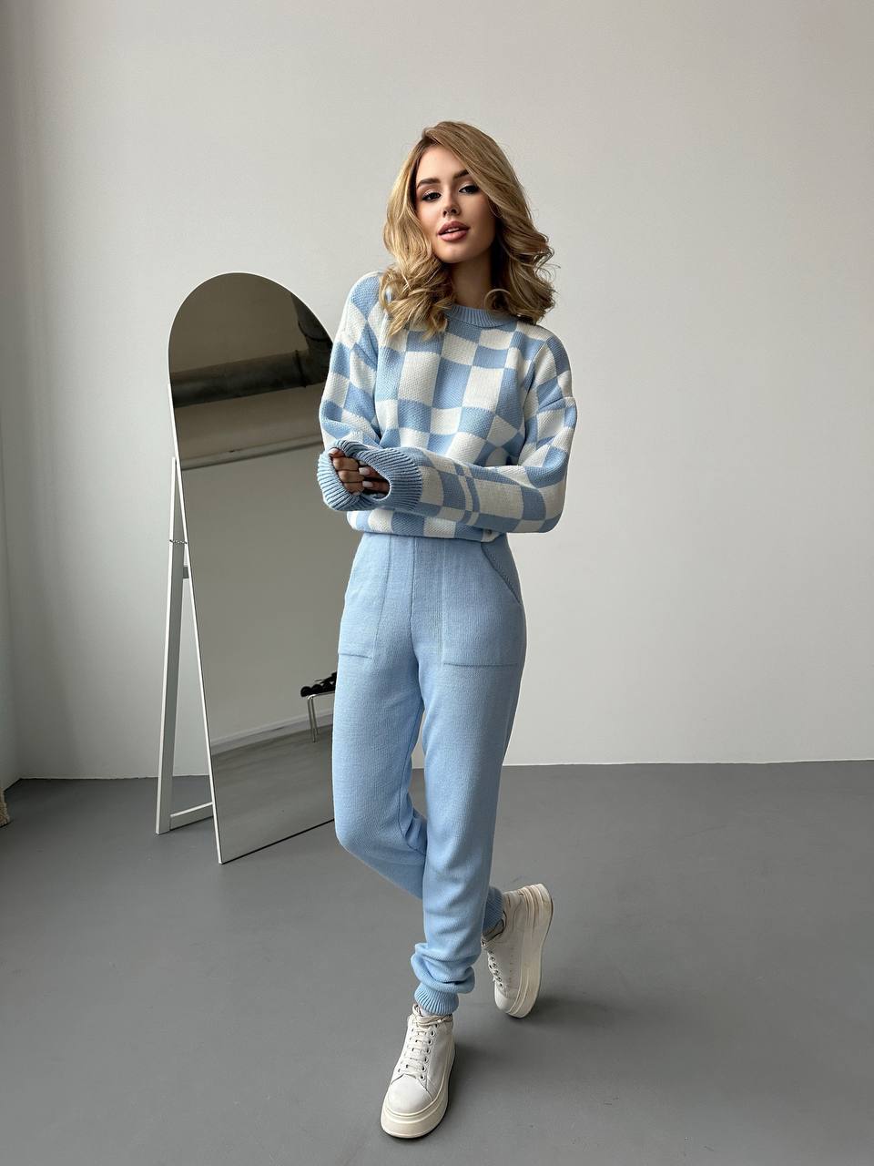 Person wearing a blue and white checkered sweater and light blue pants, standing in a minimalistic room.
