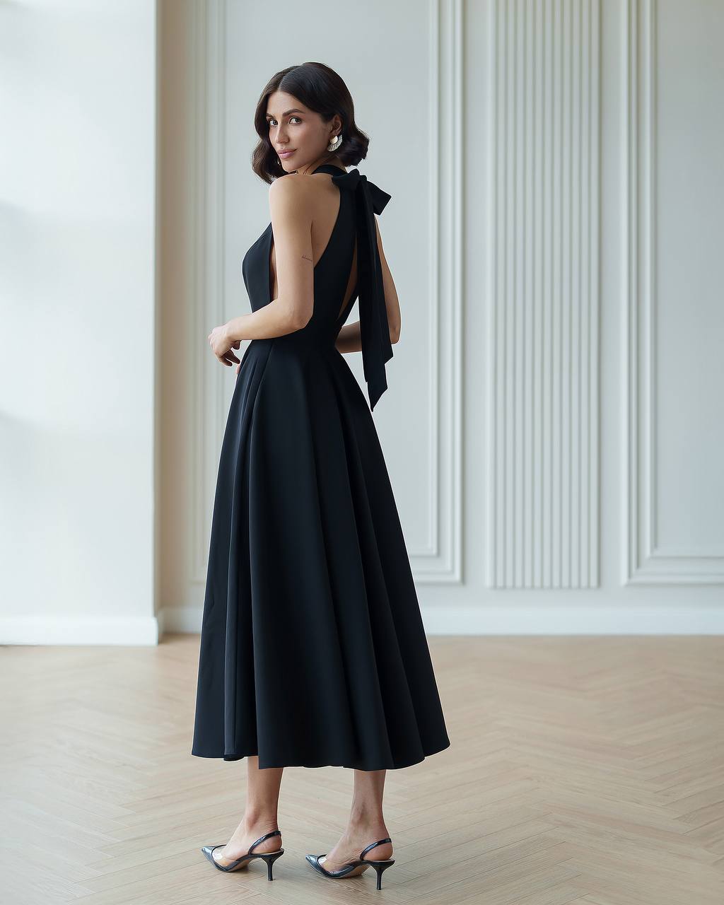 Woman wearing a black dress with a bow detail in a room with white walls and wooden floor.