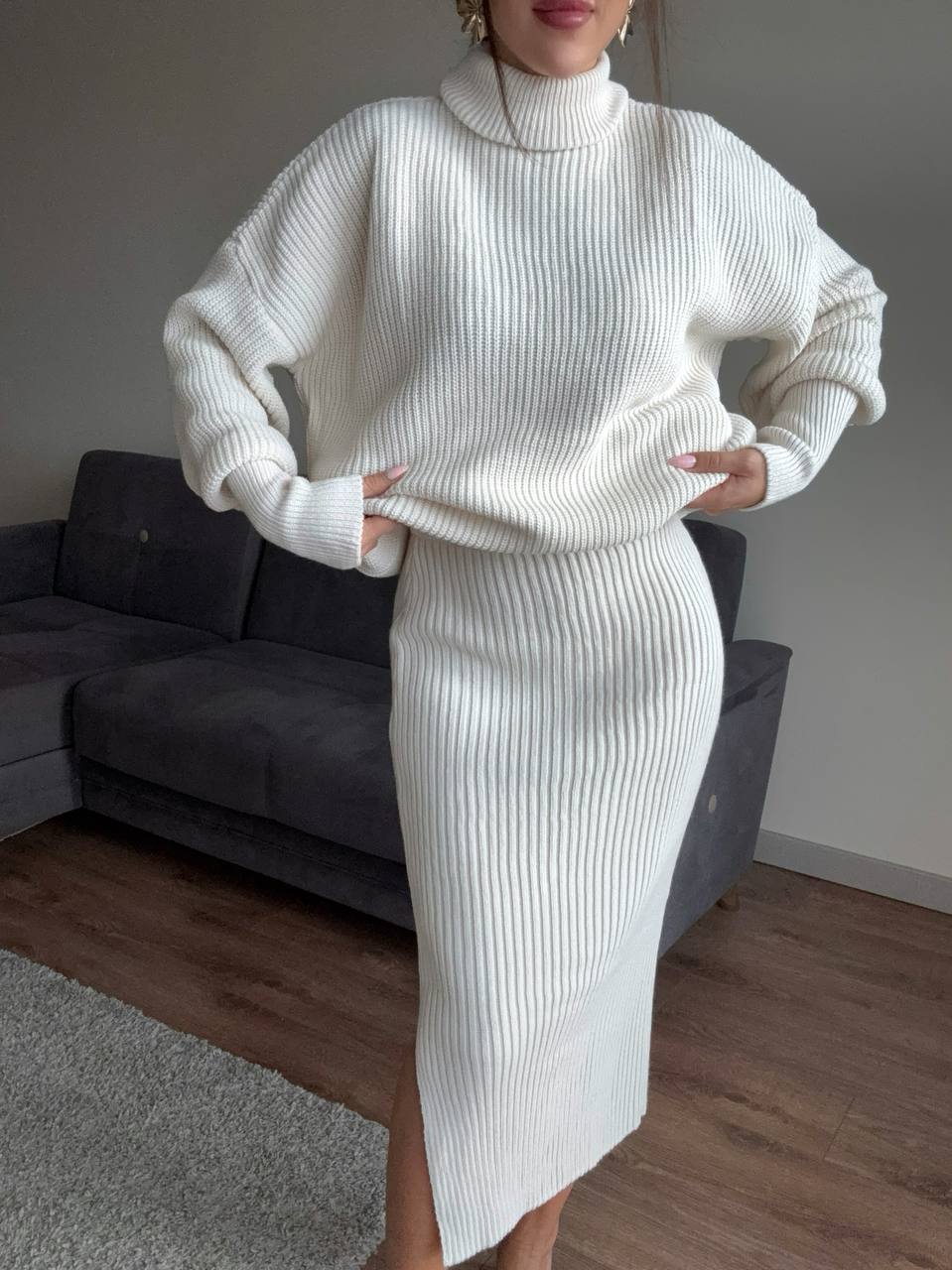 Person wearing a white ribbed sweater and skirt set in a room with a gray sofa.