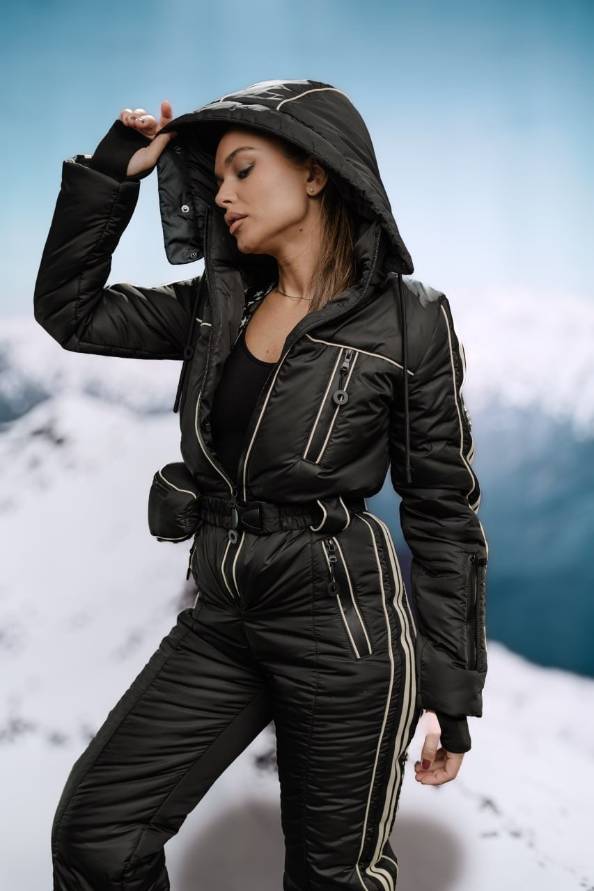 Person wearing a black snowsuit with a hood against a snowy mountain background