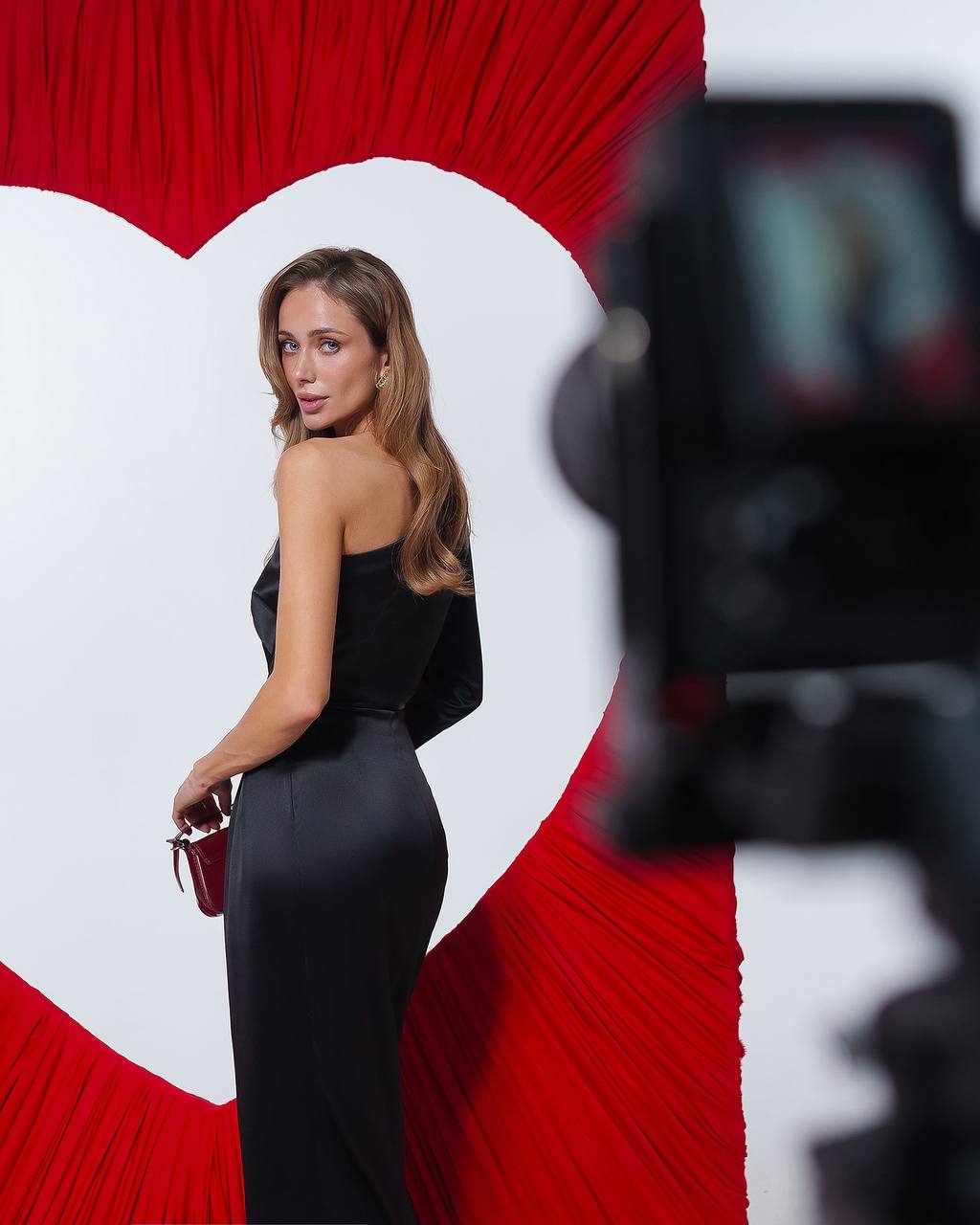 Woman in a black dress posing in front of a red heart-shaped backdrop with a camera in the foreground.