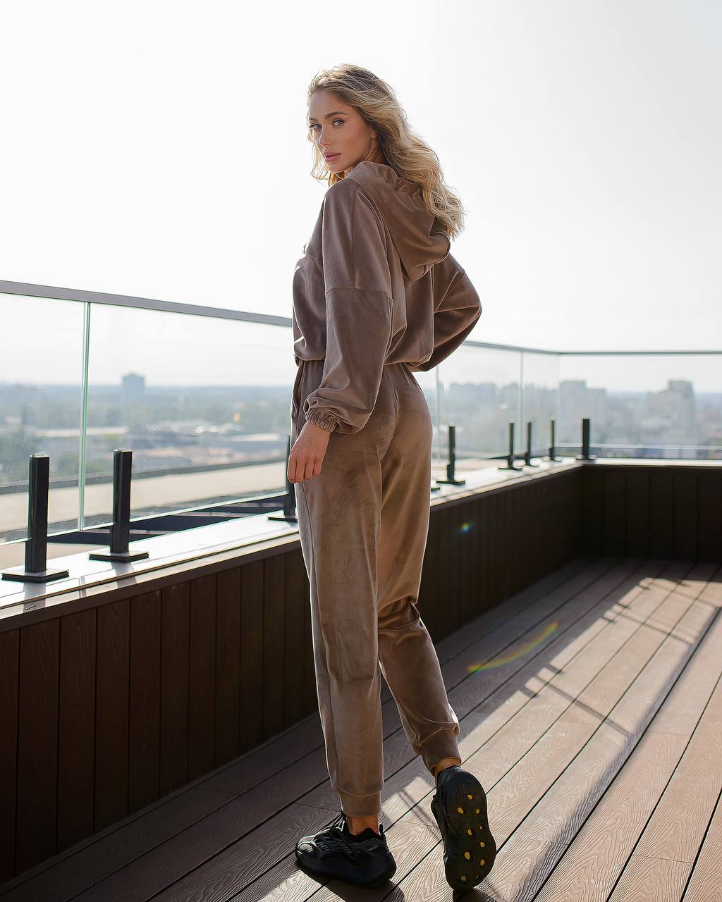 Woman in a beige tracksuit standing on a rooftop with a cityscape view.