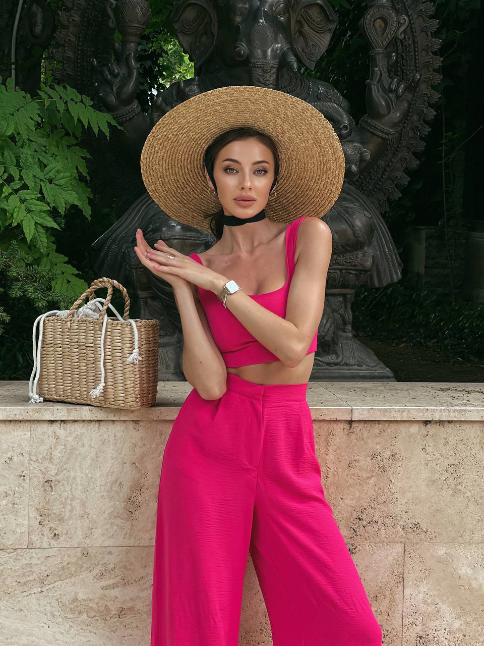 Woman in a bright pink outfit with a wide-brimmed hat standing in front of a stone wall and statue.