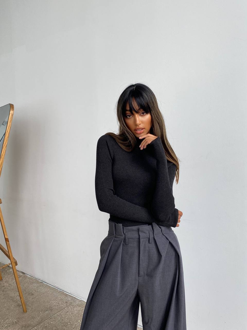 Woman wearing a black sweater and gray pants against a plain wall.