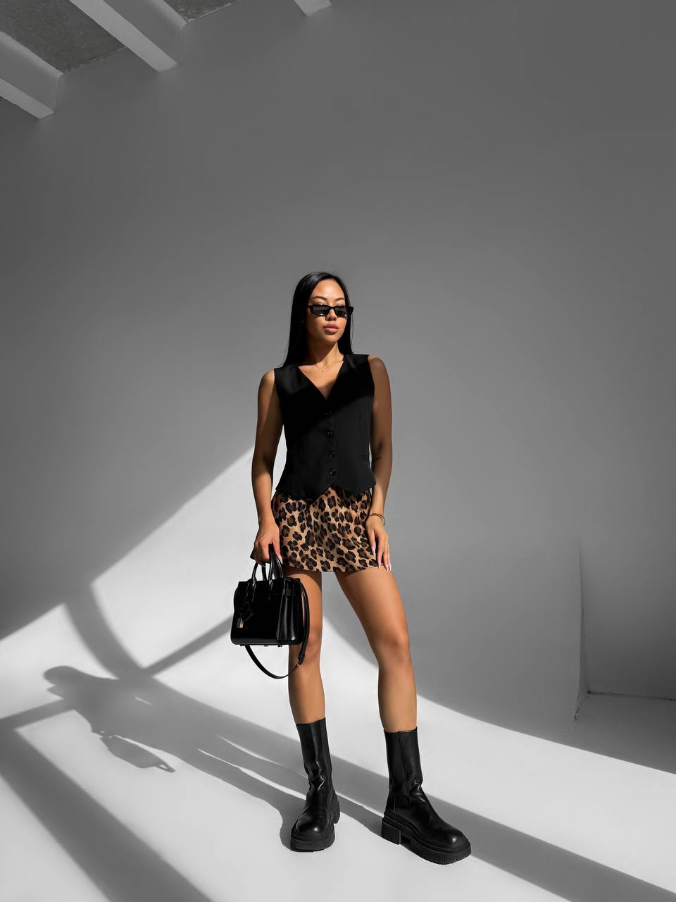 Woman in black sleeveless top, leopard print shorts, and black boots holding a black handbag against a gray background.