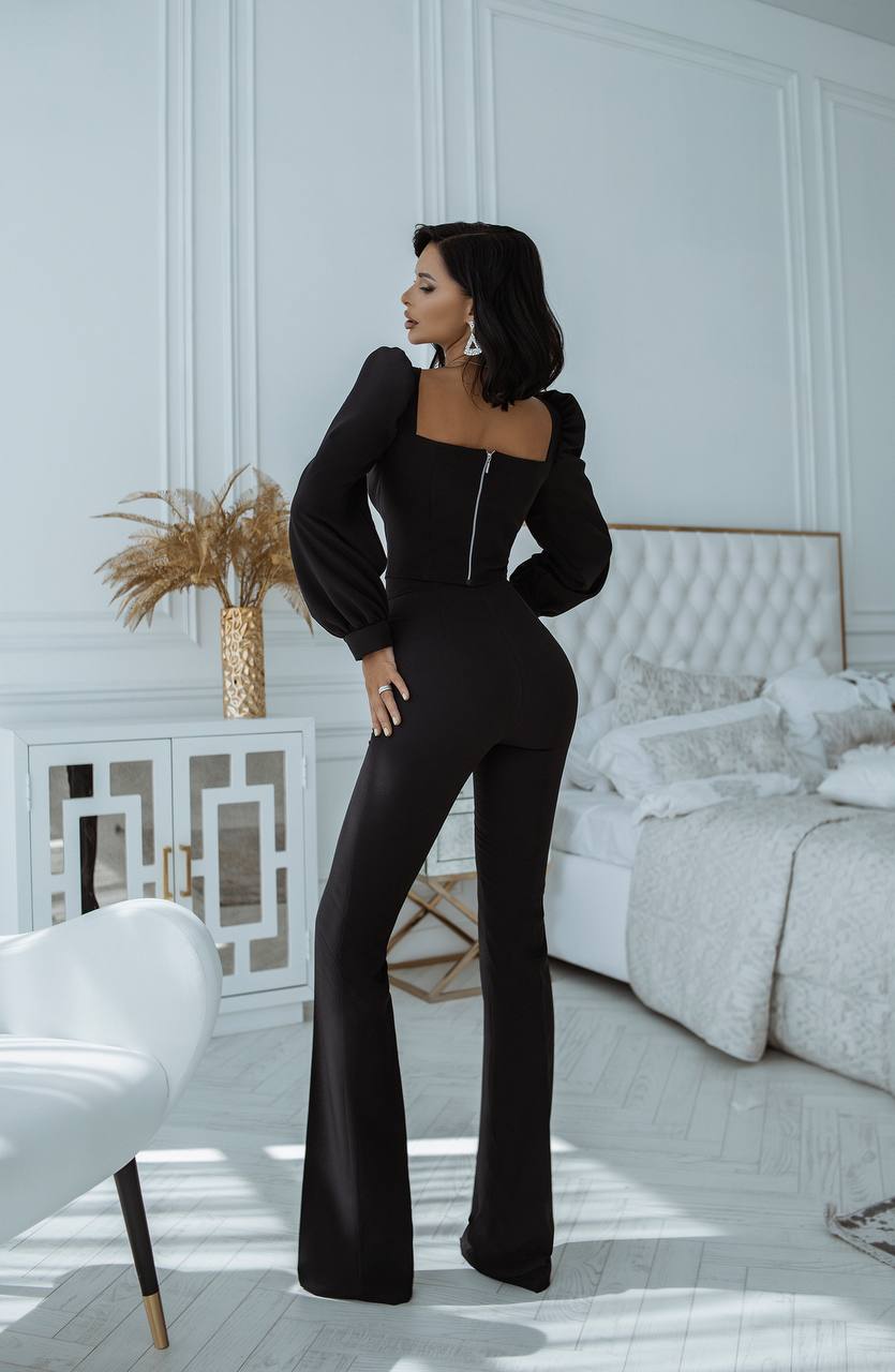 Woman in a black jumpsuit standing in a stylish bedroom.