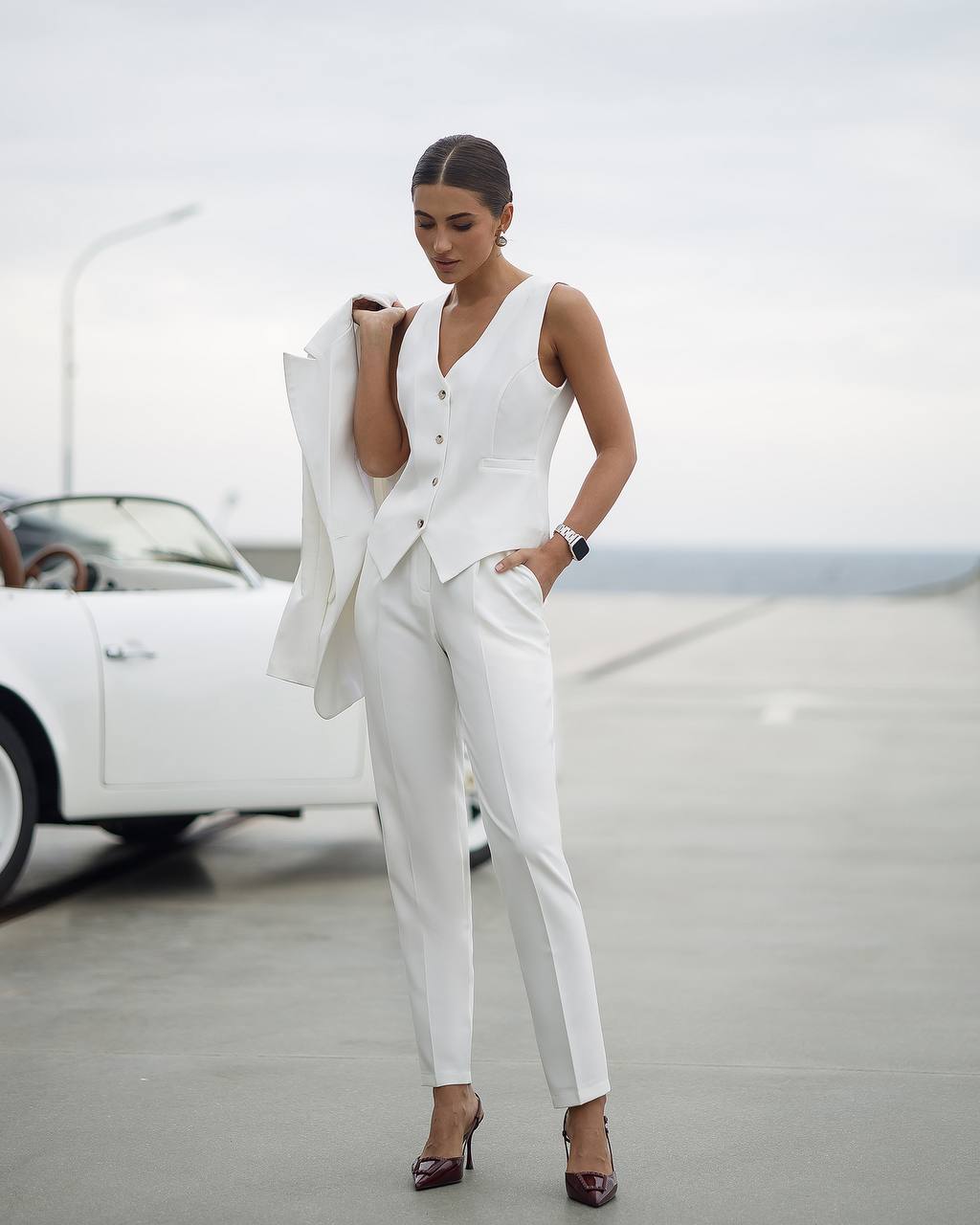 Woman in a white suit standing in front of a vintage car on a road.