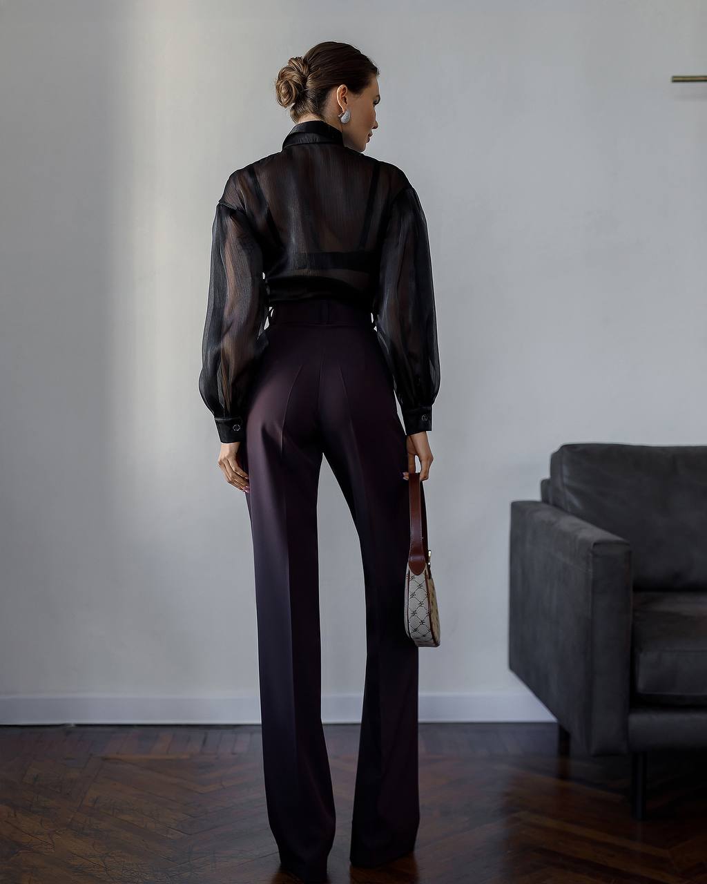 Woman in a black sheer blouse and dark pants standing in a room with a gray sofa.