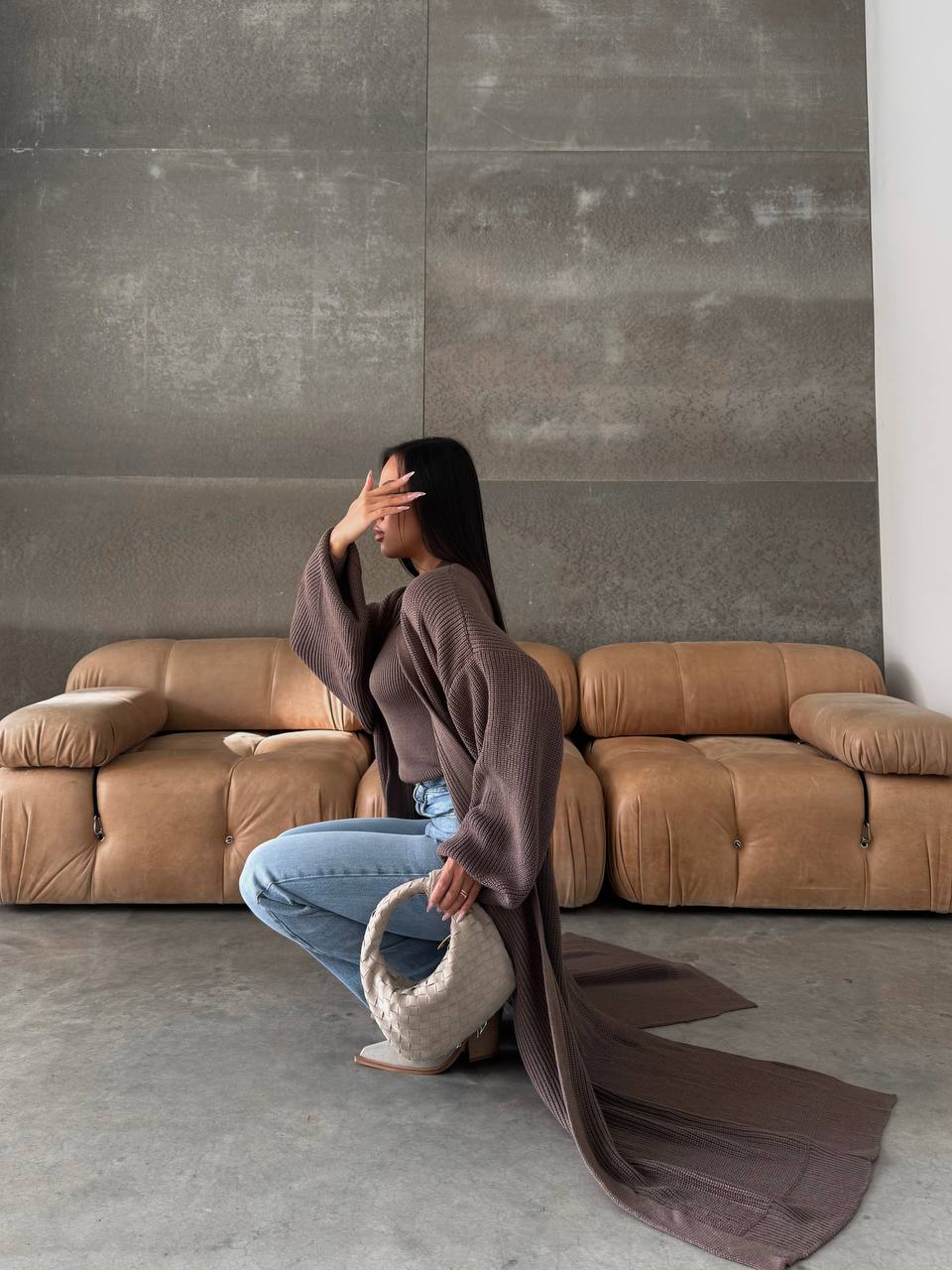 Woman in a brown sweater and jeans sitting on a beige sofa against a gray wall.