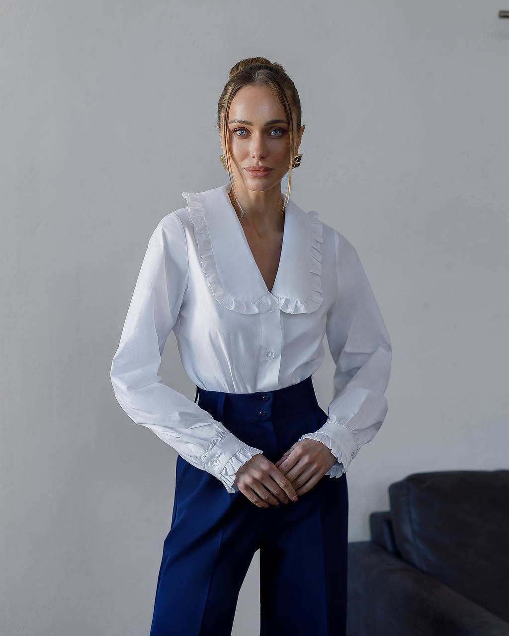 Woman wearing a white blouse with ruffled details and blue pants against a plain background