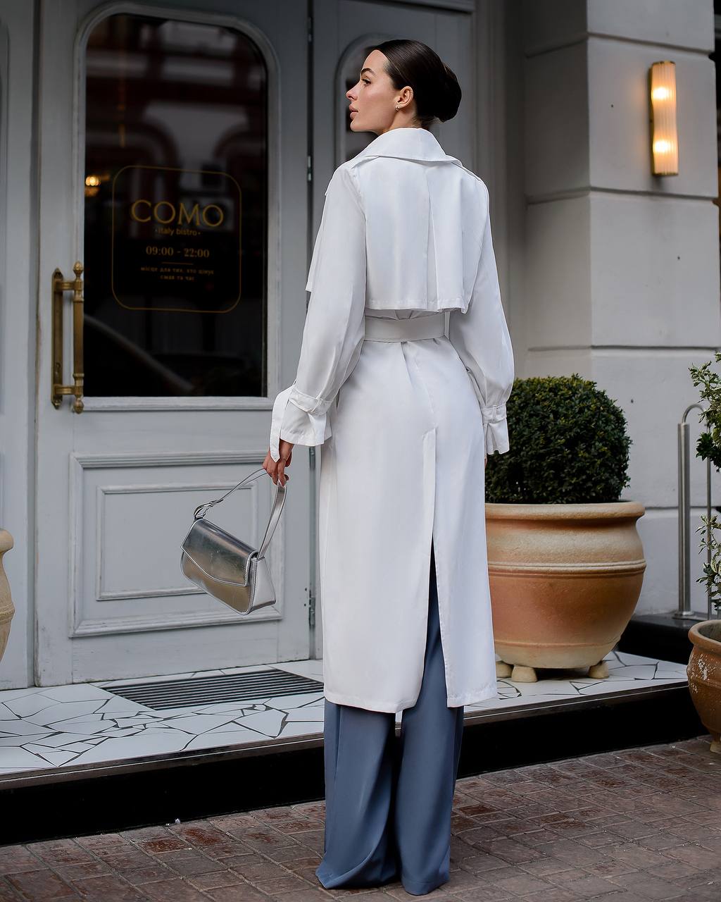 Woman in a white coat standing in front of a building entrance.