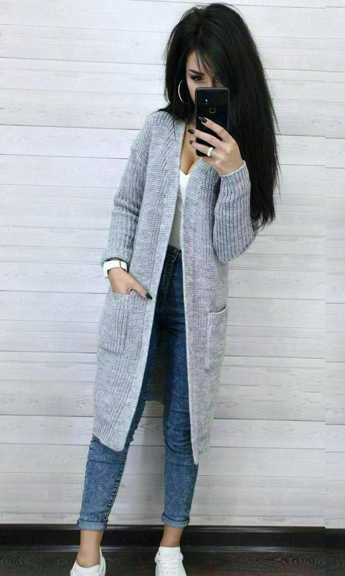 Person wearing a long gray cardigan over a white top and blue jeans, standing against a light wooden panel background.