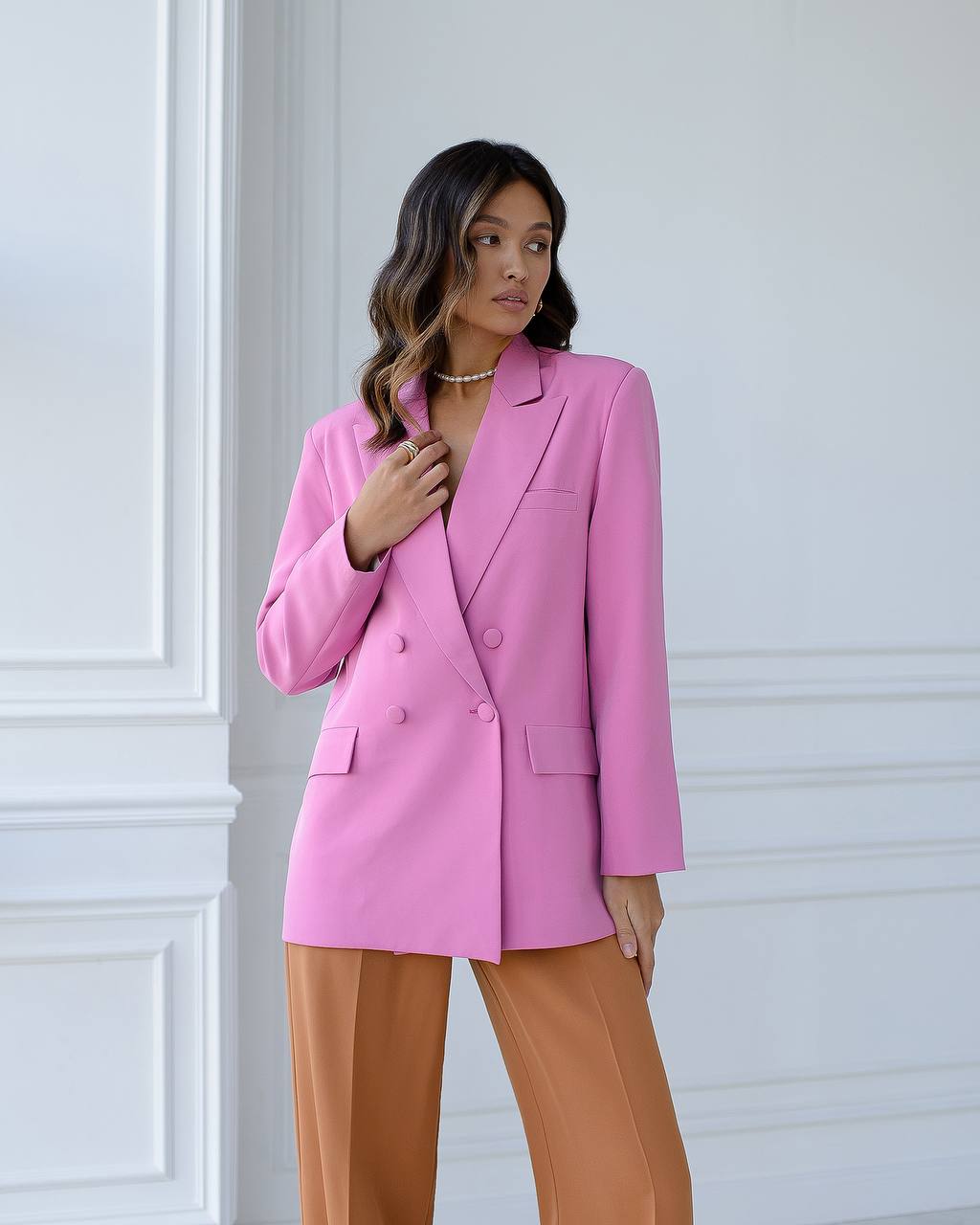 Woman wearing a pink blazer and brown pants against a white wall