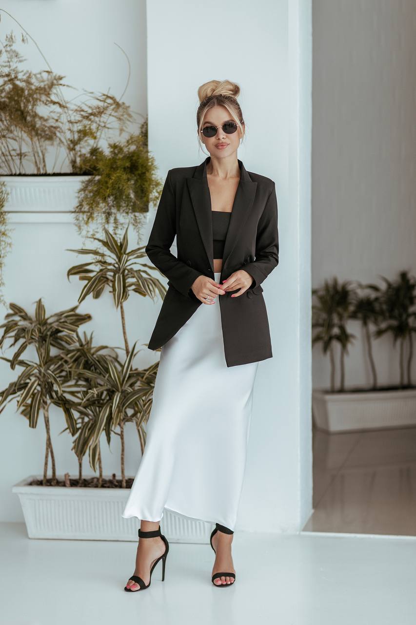 Woman in a black blazer and white dress standing indoors with plants in the background