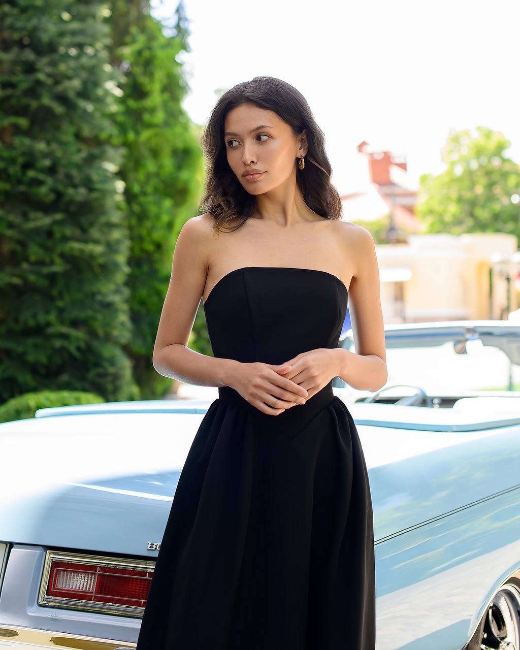 Woman in a black strapless dress standing next to a vintage car with greenery in the background