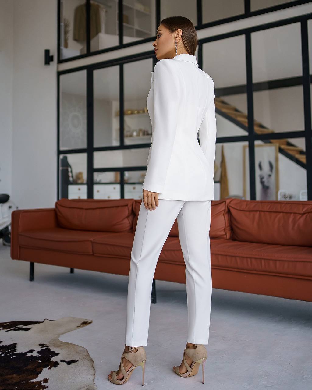 Woman in a white suit standing in a modern living room with a brown sofa.
