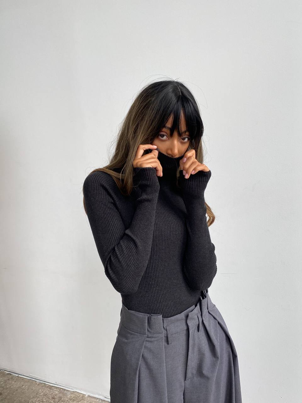 Person wearing a black turtleneck and gray pants against a white wall