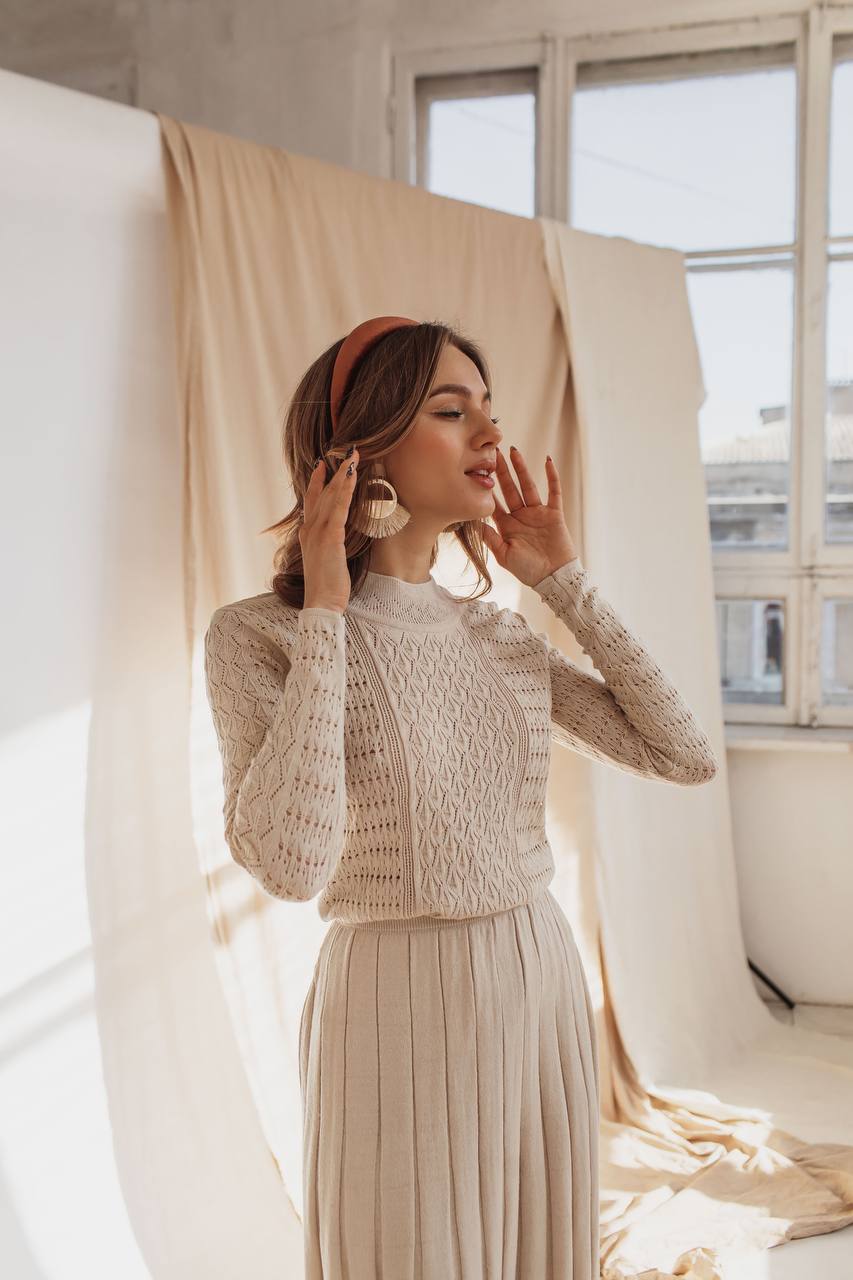 Woman wearing a beige crochet top and pleated skirt in a softly lit room.