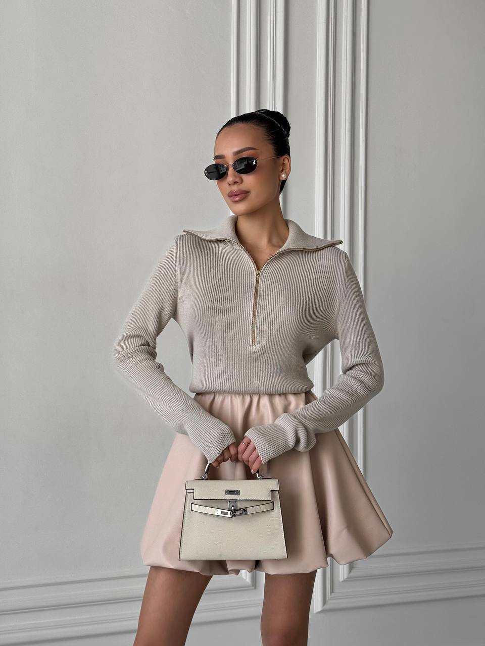 Woman in a beige sweater and skirt holding a white handbag against a gray wall.