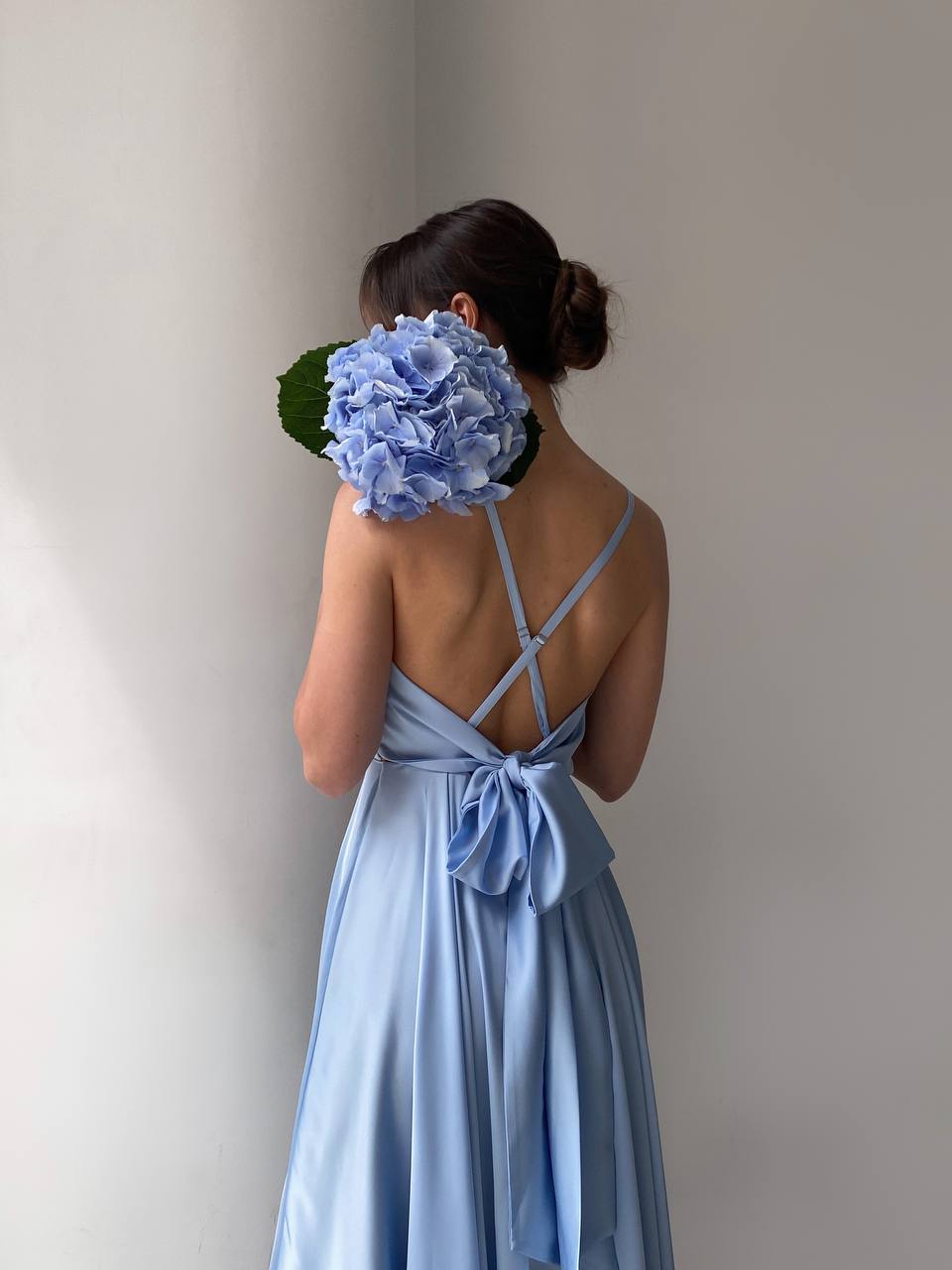 Woman in a light blue dress holding a large blue flower against a plain background
