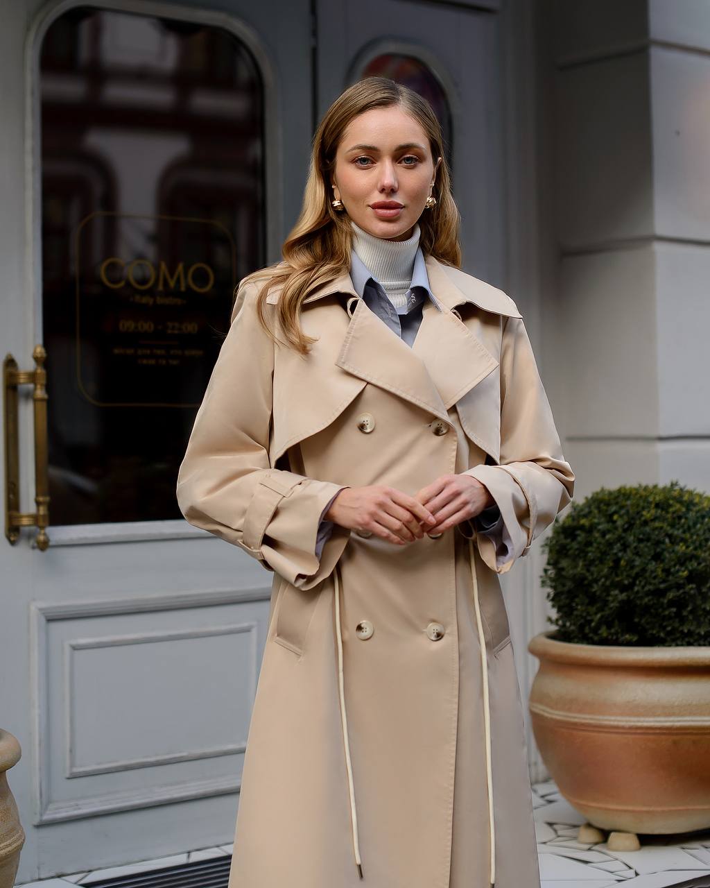 Woman wearing a beige trench coat standing in front of a building with 'COMO' branding.