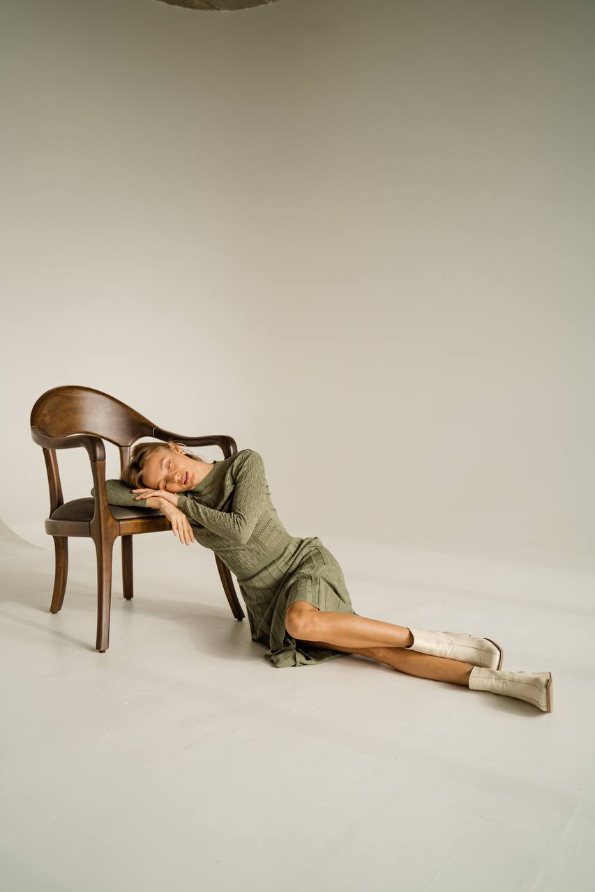 Woman in a green dress and white boots lying on the floor next to a wooden chair against a plain background