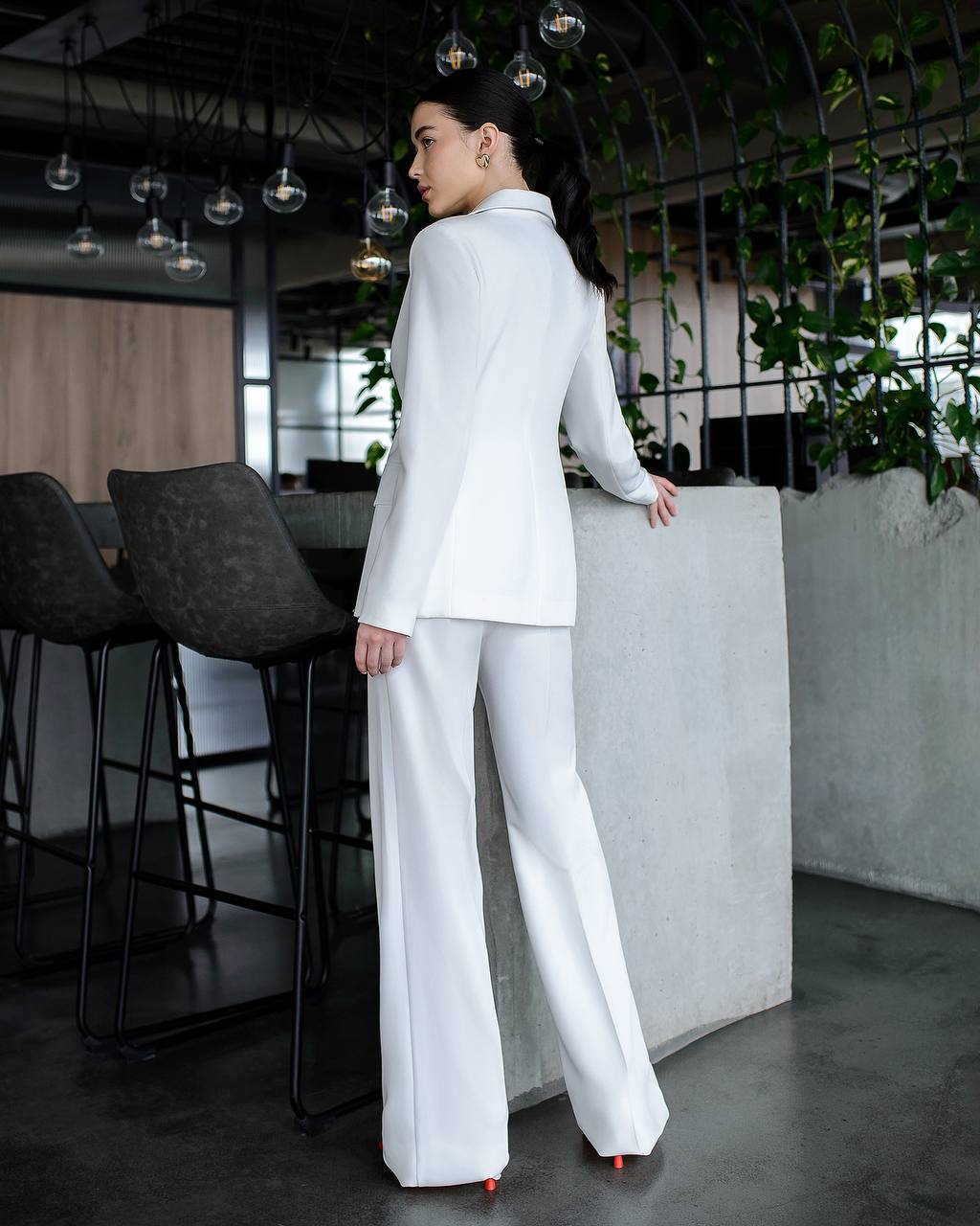 Person wearing a white suit standing in an indoor setting with plants and chairs.