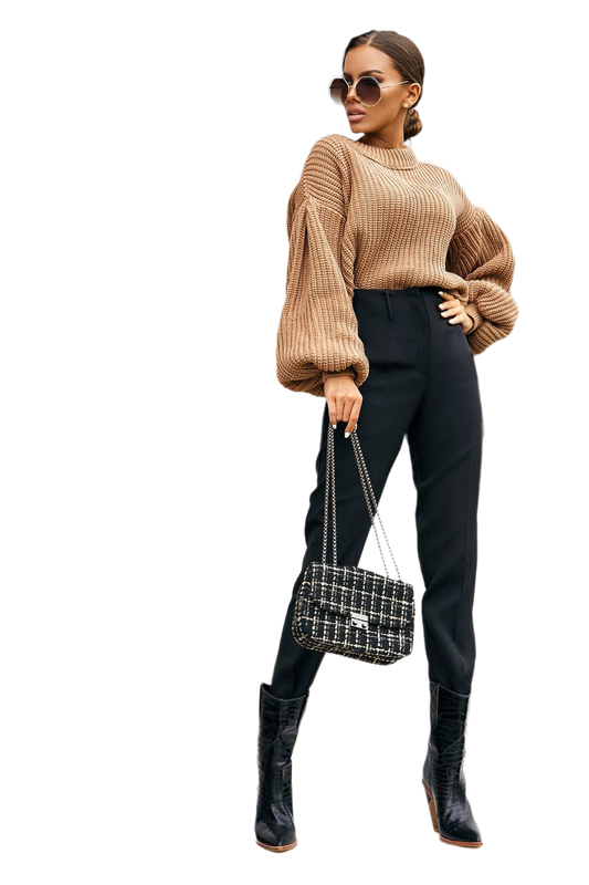 Woman wearing a beige sweater, black pants, and black boots holding a patterned handbag on a white background
