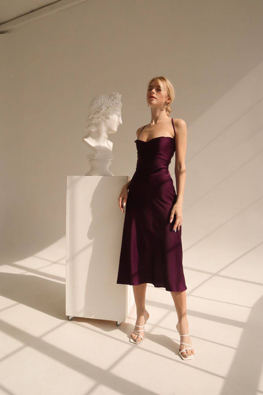 Woman in a dark purple dress standing next to a classical sculpture in a well-lit room.
