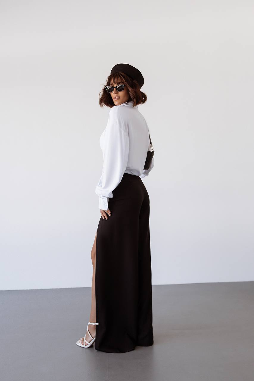 Woman wearing a white blouse and black pants with a high slit, standing against a plain background.