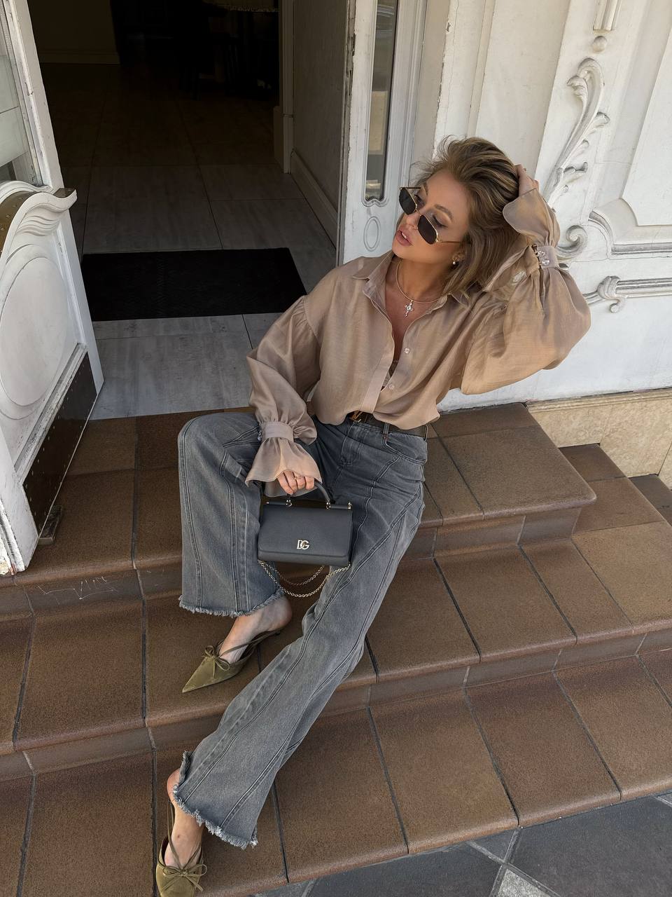 Woman sitting on steps wearing a beige blouse and blue jeans, holding a black handbag.
