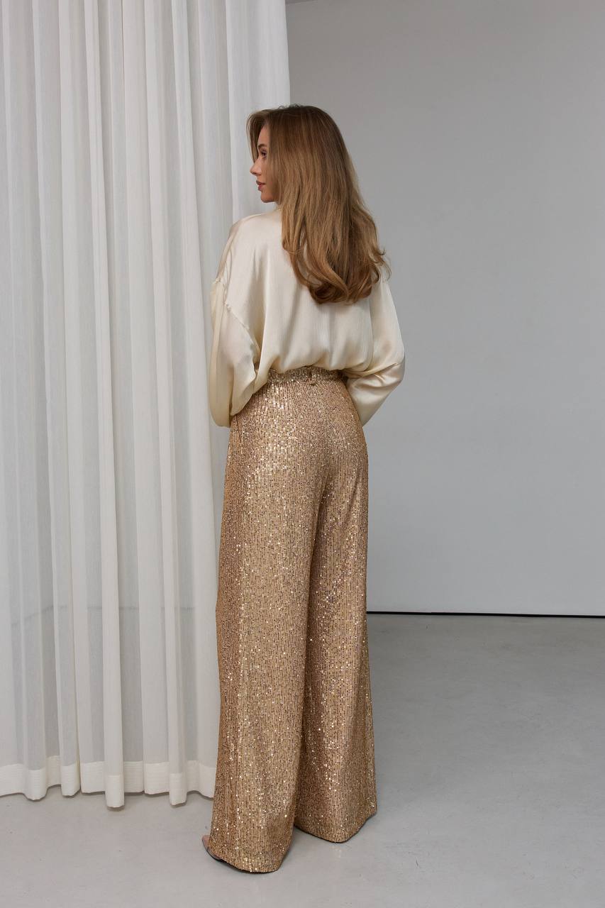 Woman wearing a white blouse and gold sequin pants standing in front of a white curtain.
