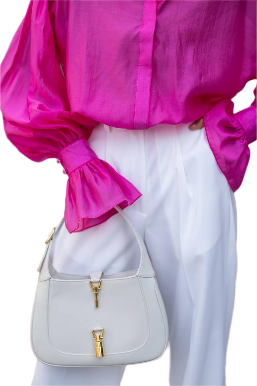 Person wearing a bright pink blouse and white pants holding a light gray handbag with gold accents.
