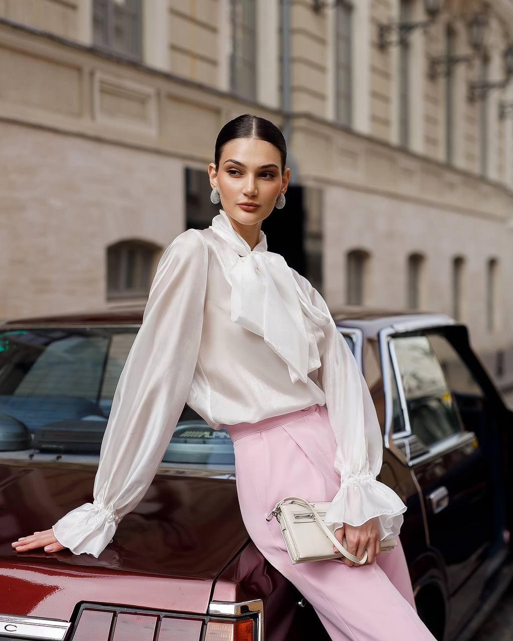 Woman in a white blouse and pink pants standing next to a vintage car with a building in the background