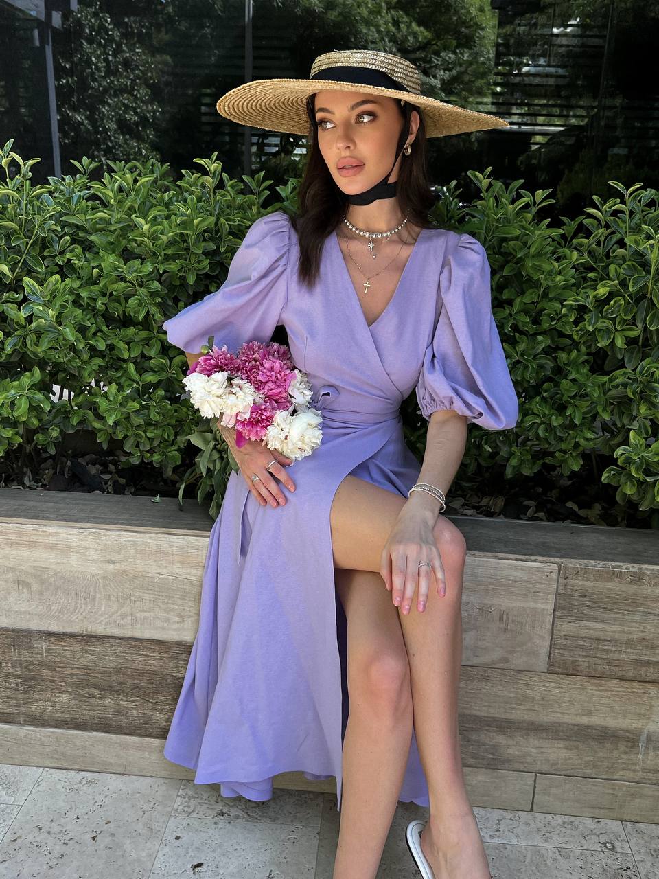 Woman in a lavender dress and wide-brimmed hat holding flowers, sitting on a stone ledge.