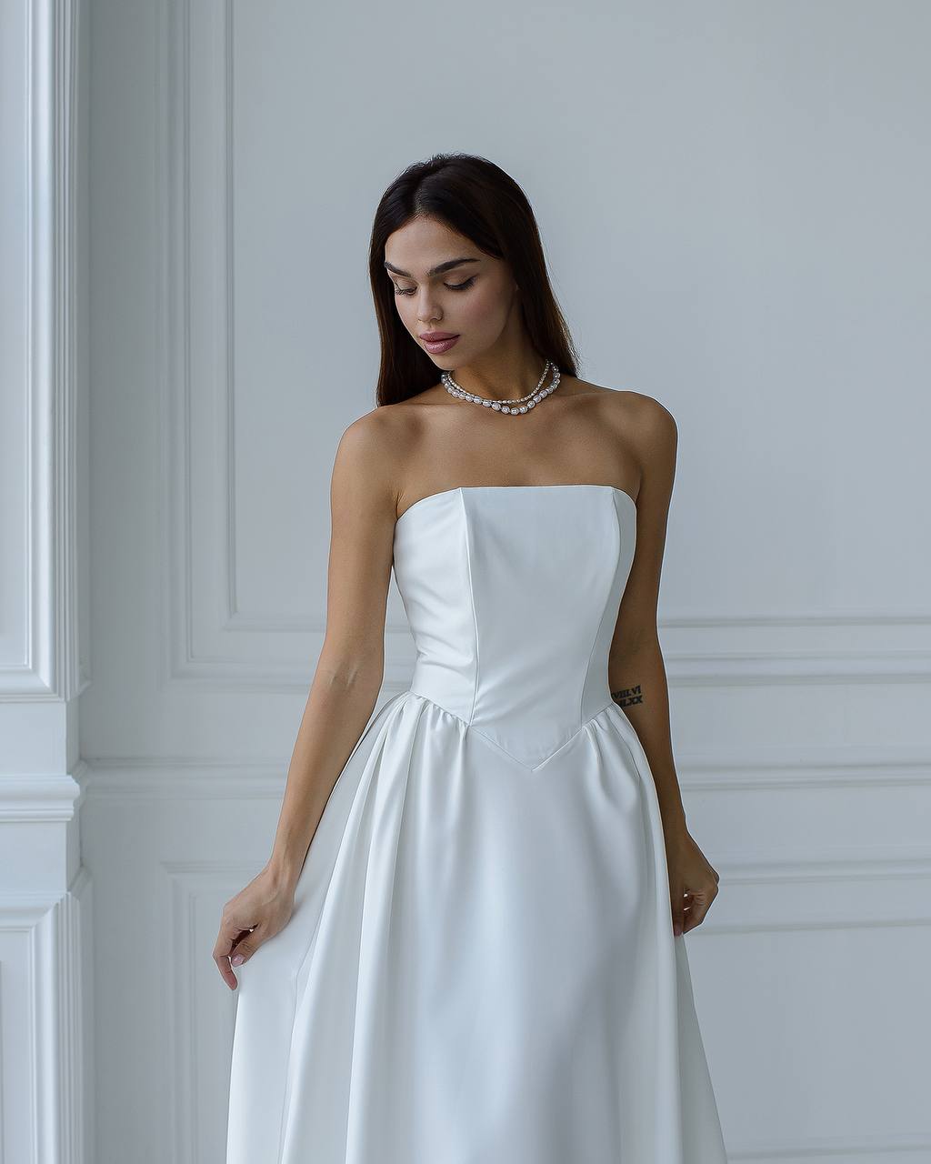 Woman wearing a strapless white wedding dress against a white wall.