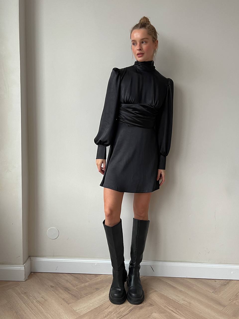 Person wearing a black dress and boots against a plain wall.