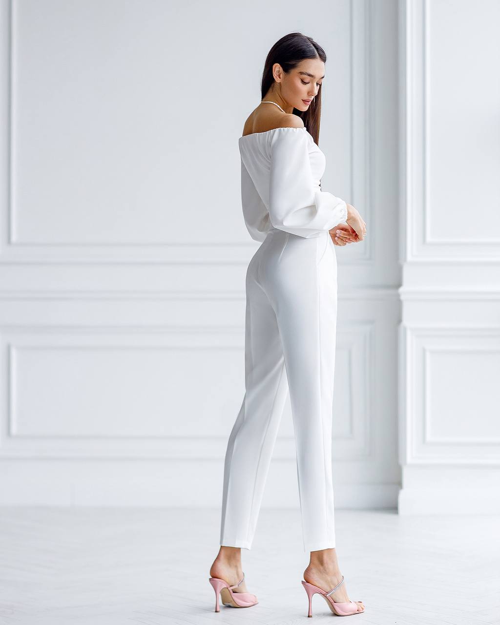 Woman wearing a white off-shoulder top and pants against a white wall.