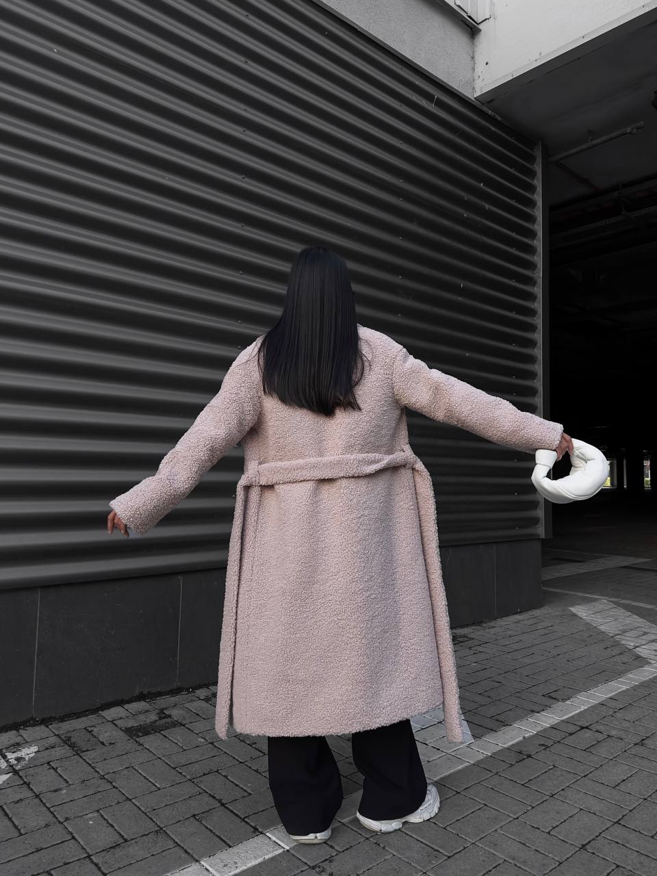 Person wearing a long coat with a belt, standing on a street with a building in the background.